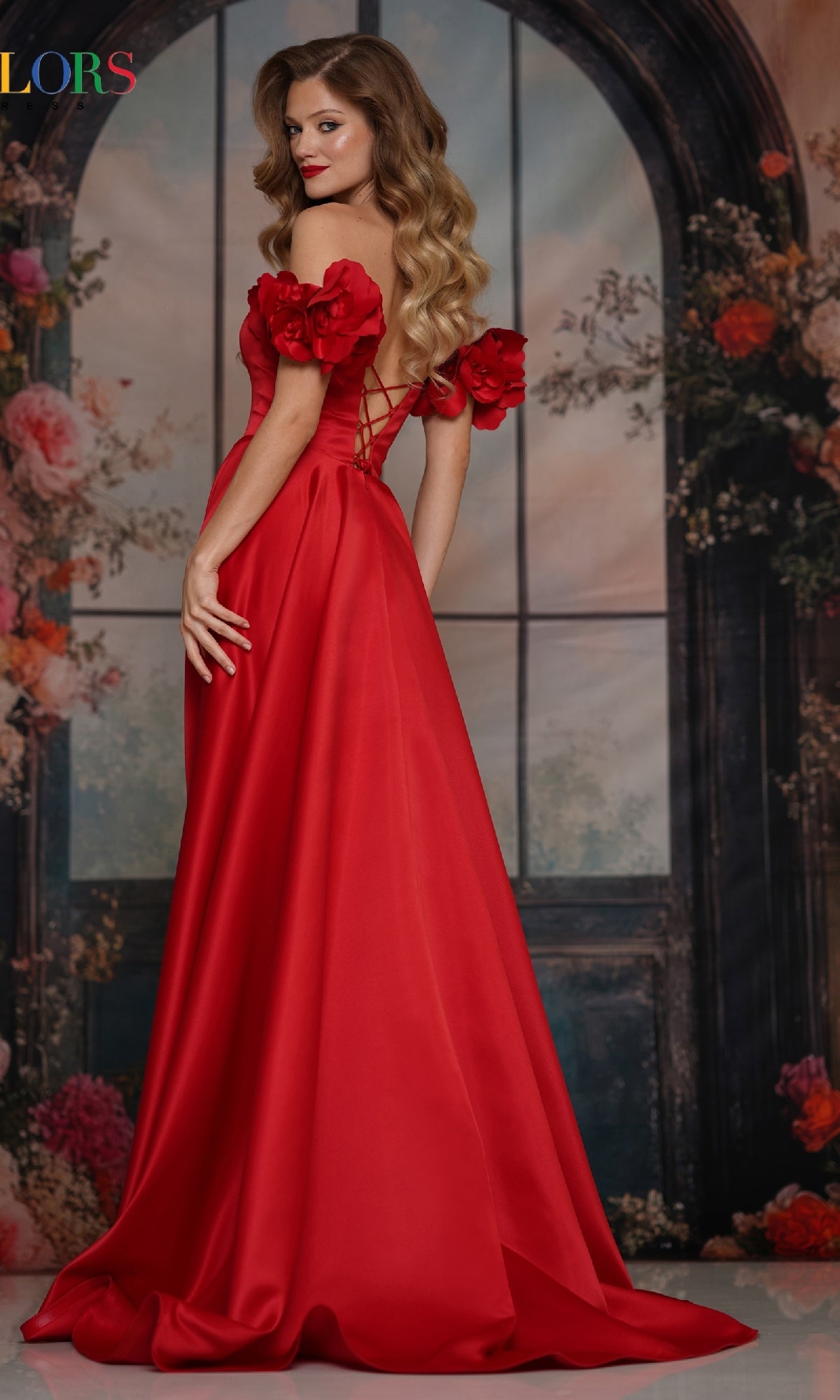 A woman with long wavy hair wears the Off-Shoulder Satin Prom Gown 3744 by Colors Dress, featuring floral details and a lace-up back, standing before a flower-adorned arched window backdrop.